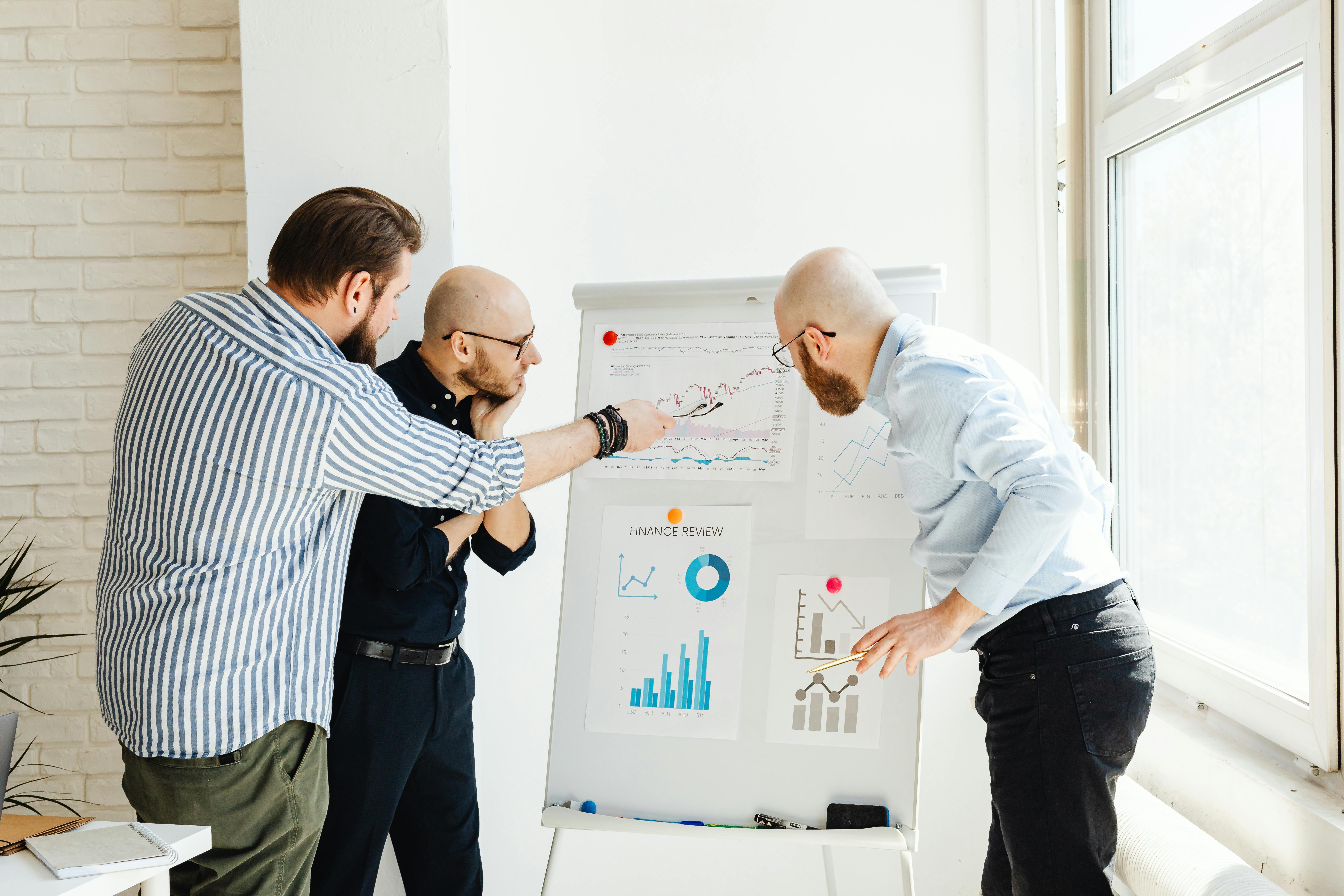Three colleagues in a bright office discuss financial charts on a whiteboard.